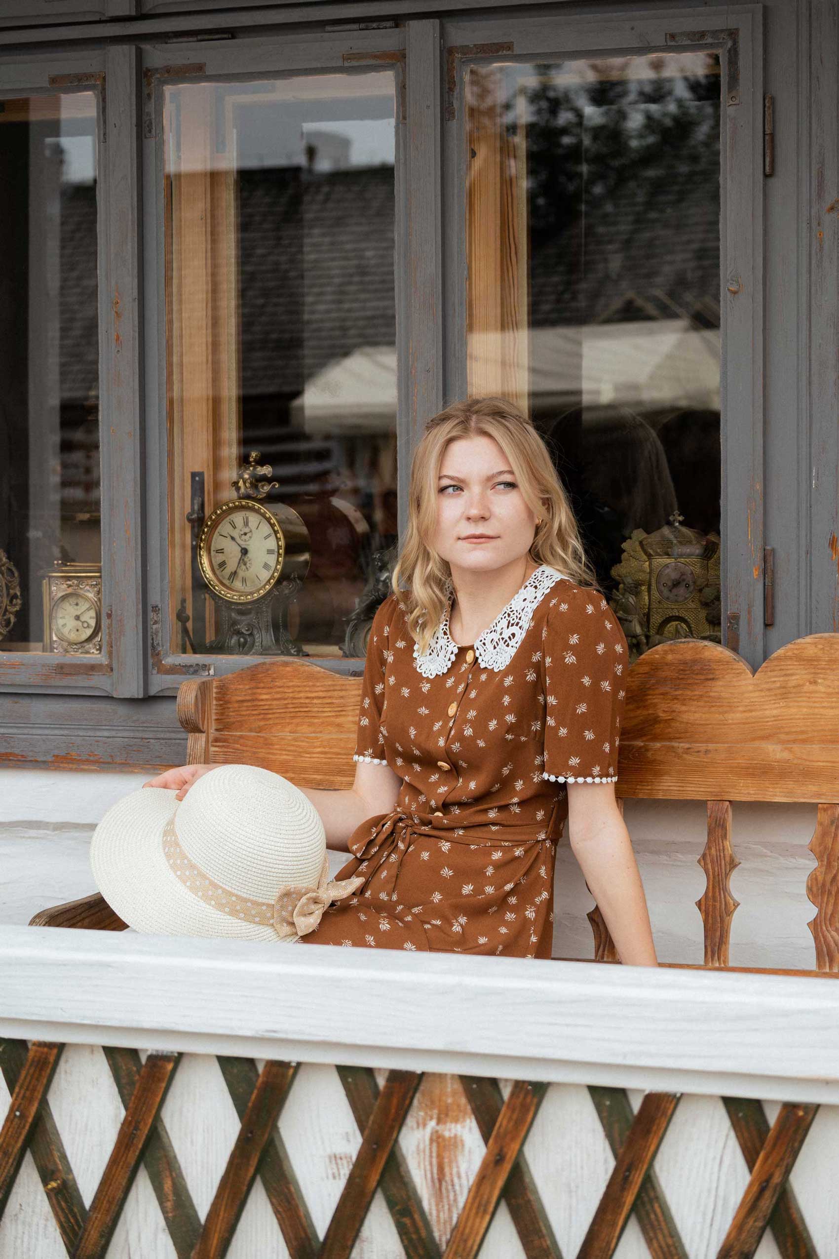 Blond dziewczyna w brązowej sukience z kapeluszem siedzi na ławce przed białym domem zegarmistrza. W oknie widać zegary