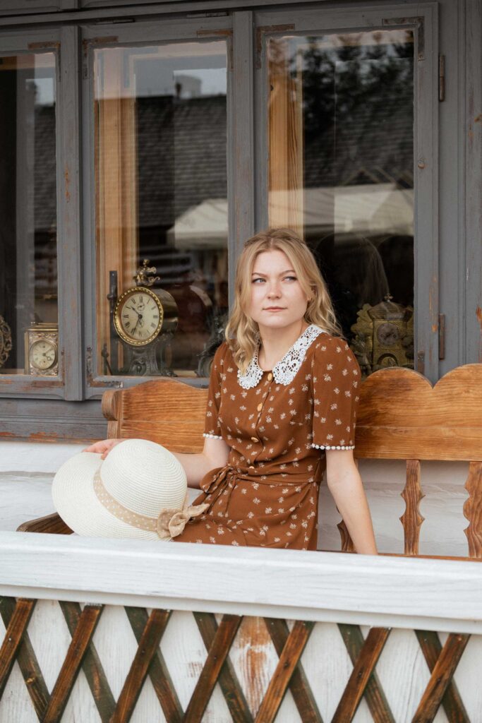 Blond dziewczyna w brązowej sukience z kapeluszem siedzi na ławce przed białym domem zegarmistrza. W oknie widać zegary