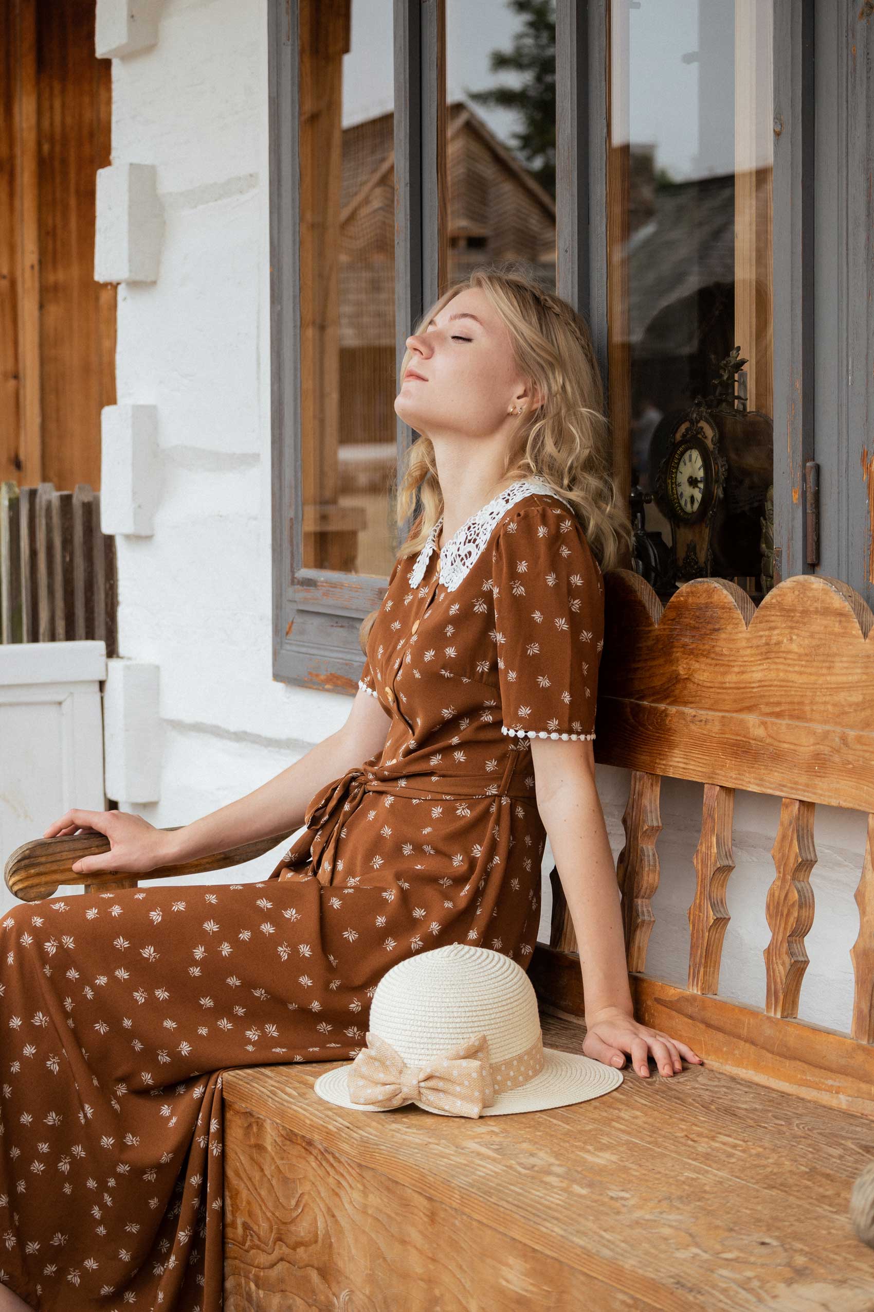 Blond dziewczyna w brązowej sukience z kapeluszem siedzi na ławce przed białym domem zegarmistrza. W oknie widać zegary