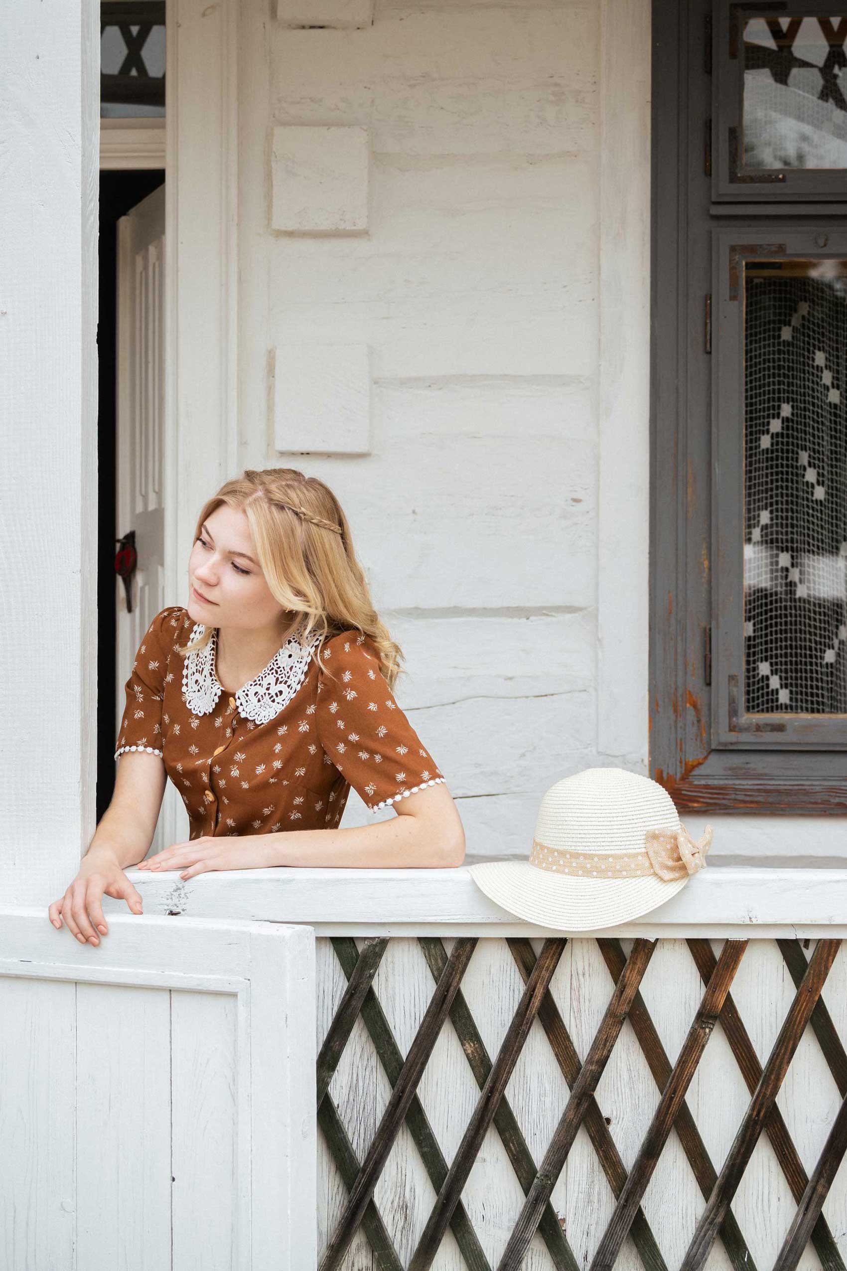 Blond dziewczyna w brązowej sukience z kapeluszem wygląda przez balustradę przed białym domem