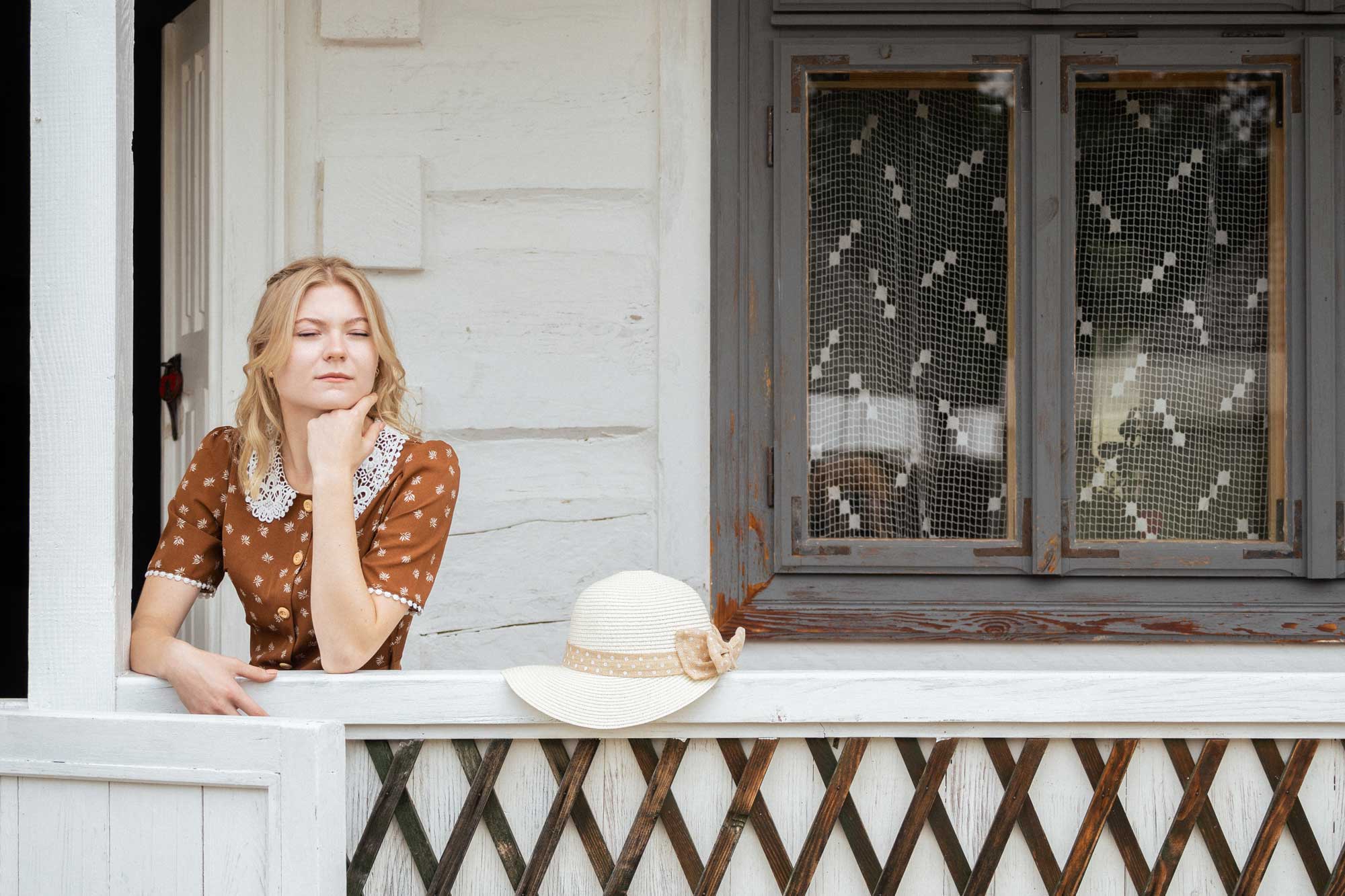 Blond dziewczyna w brązowej sukience z kapeluszem wygląda przez balustradę przed białym domem