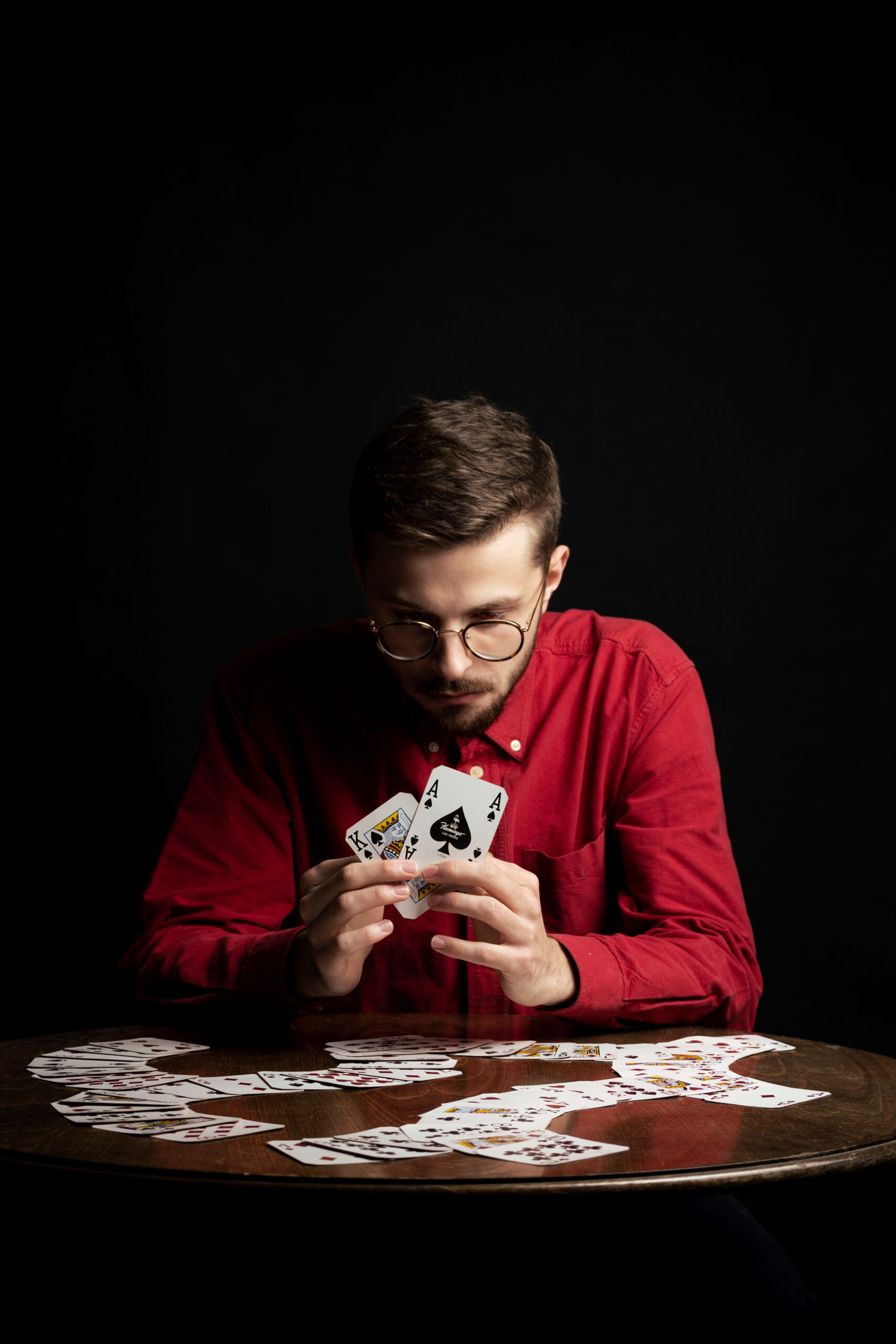 Mężczyzna w czerwonej koszuli w okularach gra na stole w karty, które są czerwone. W dłoni trzyma wachlarz z kart.