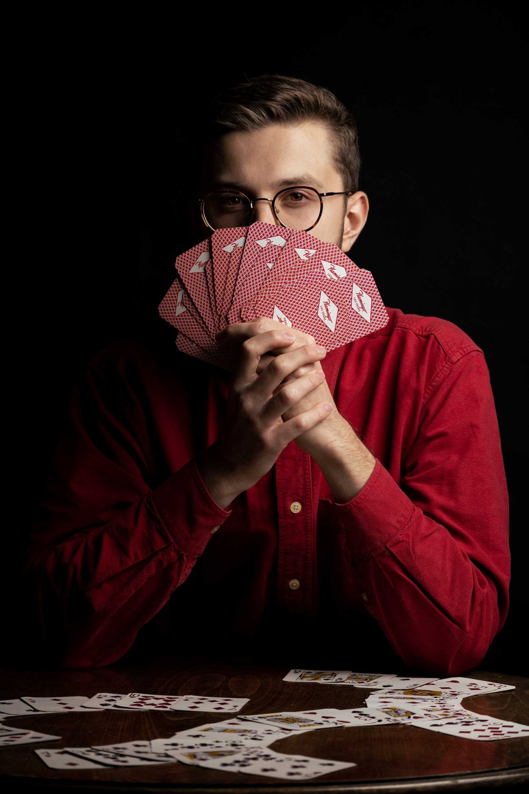 Mężczyzna w czerwonej koszuli w okularach gra na stole w karty, które są czerwone. W dłoni trzyma wachlarz z kart