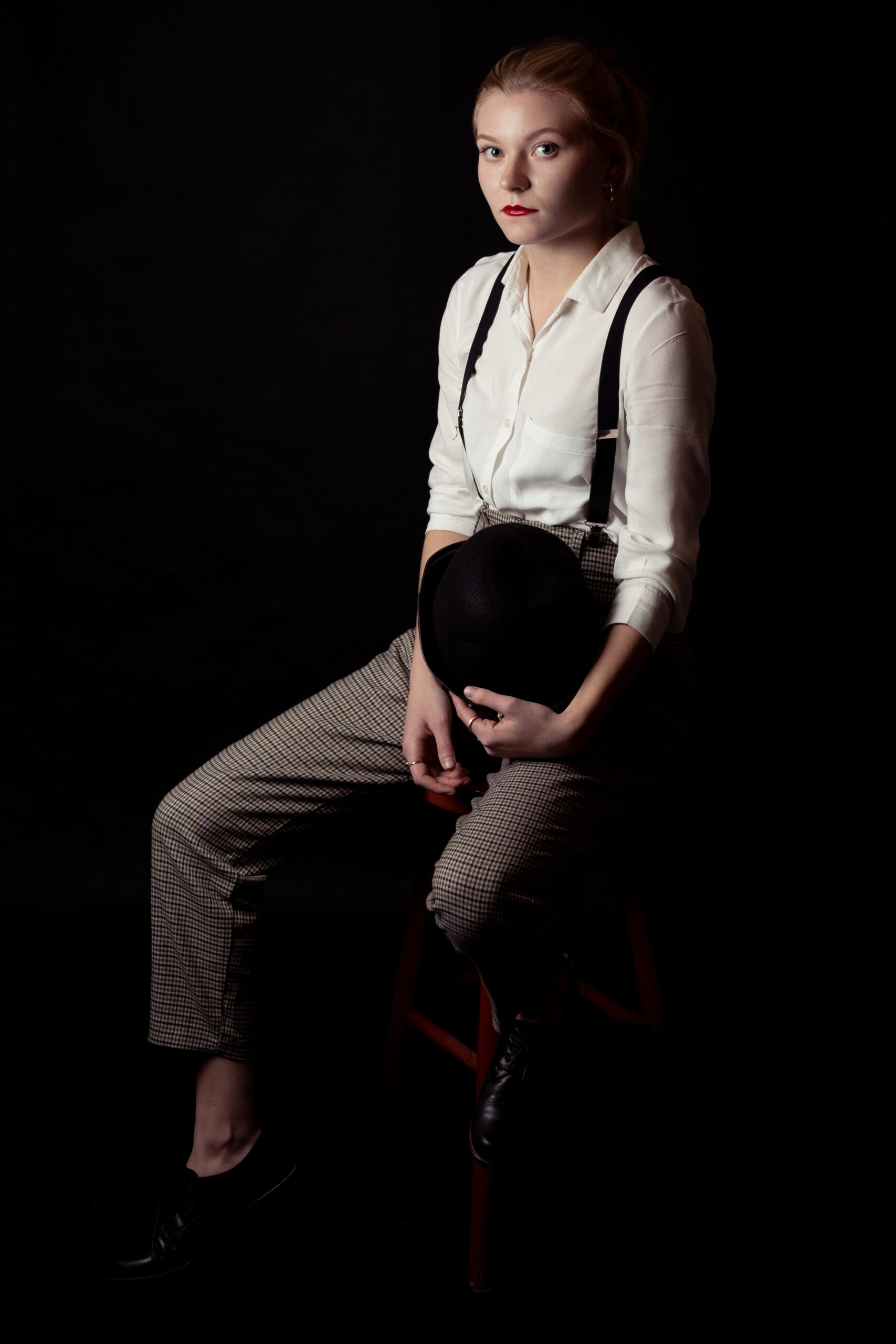 kobieta w białej koszuli, szelkach, spodniach w kratę, z czerwonymi ustami z maską i melonikiem w ręce