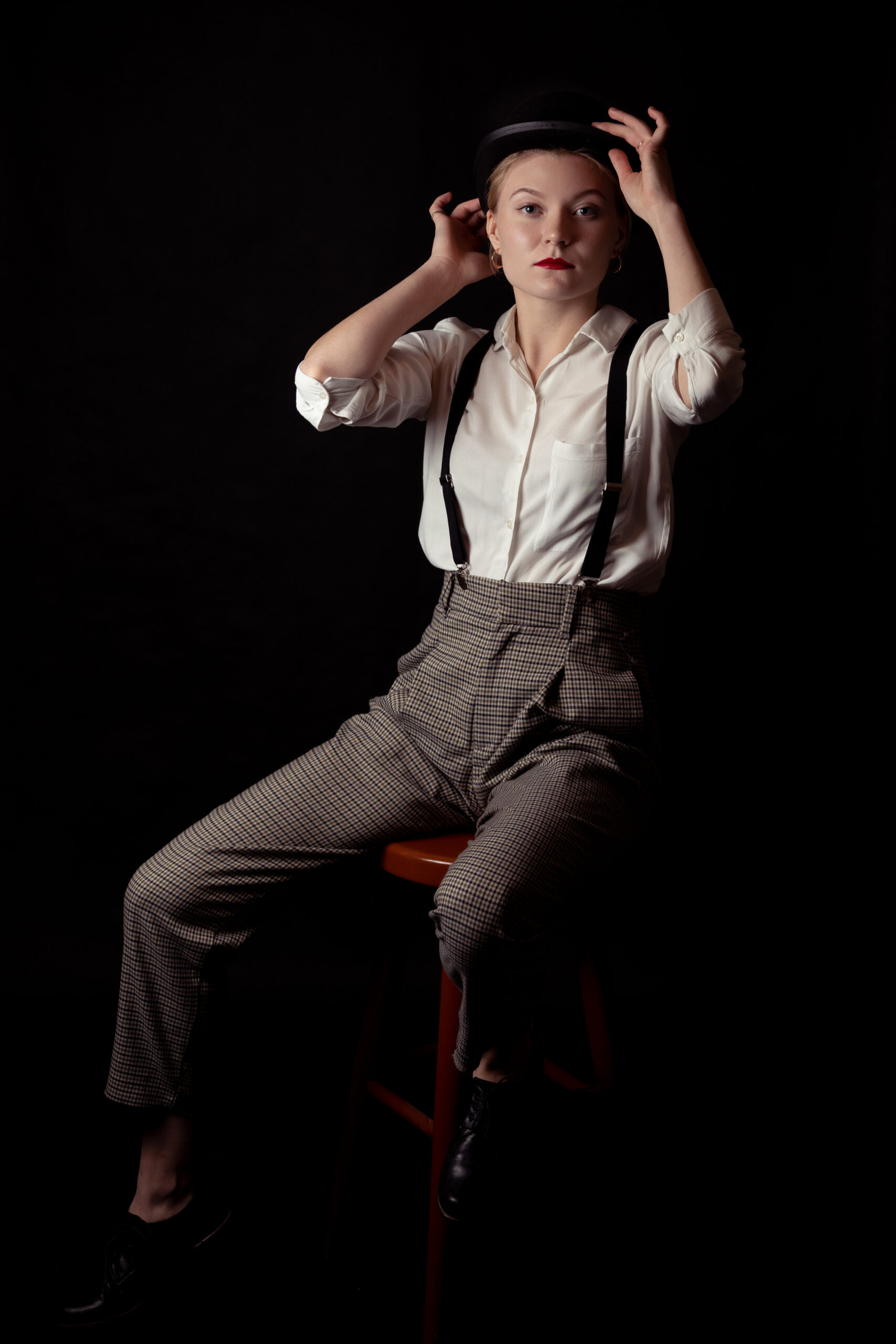 kobieta w białej koszuli, szelkach, spodniach w kratę, z czerwonymi ustami z maską i melonikiem w ręce