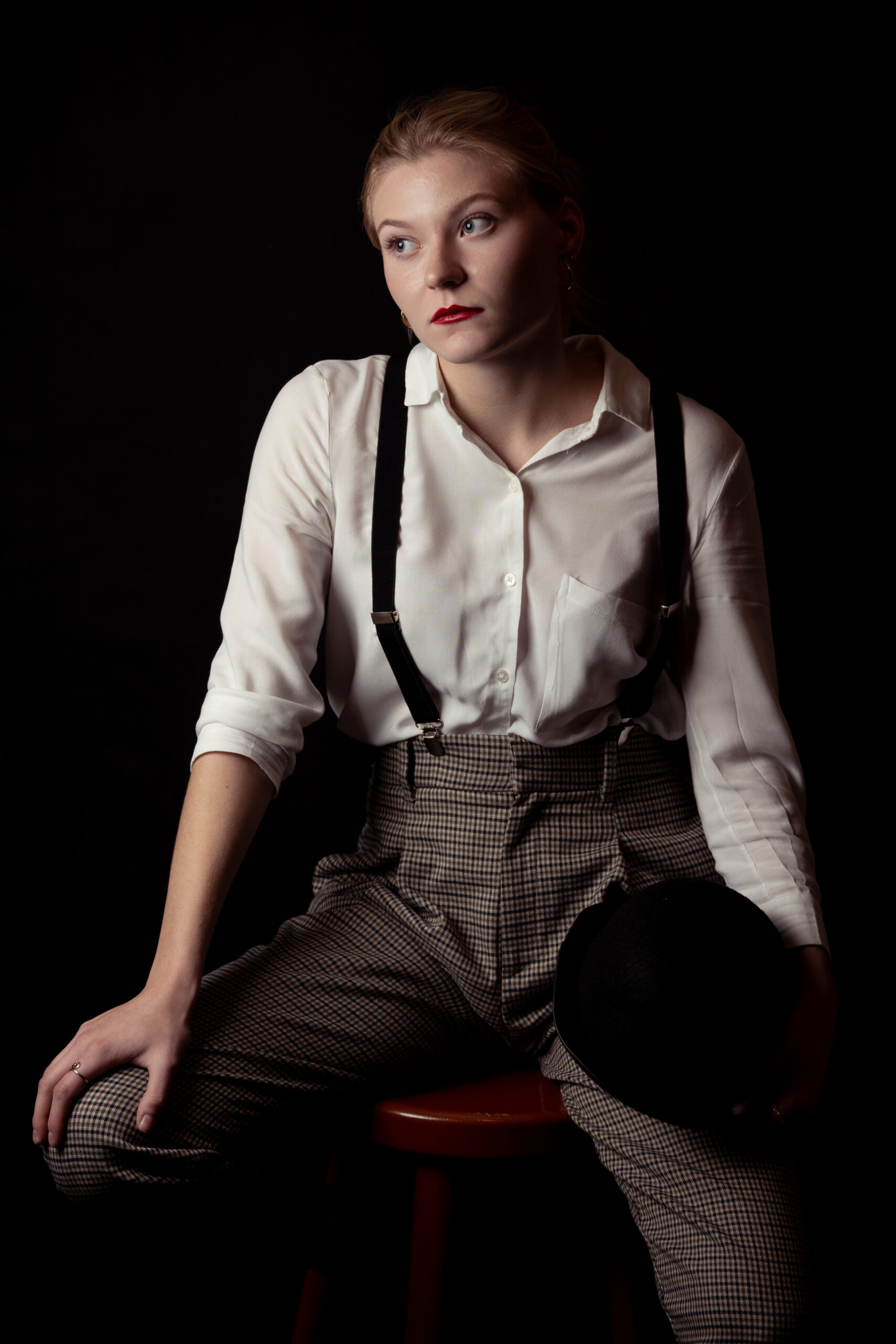 kobieta w białej koszuli, szelkach, spodniach w kratę, z czerwonymi ustami z maską i melonikiem w ręce