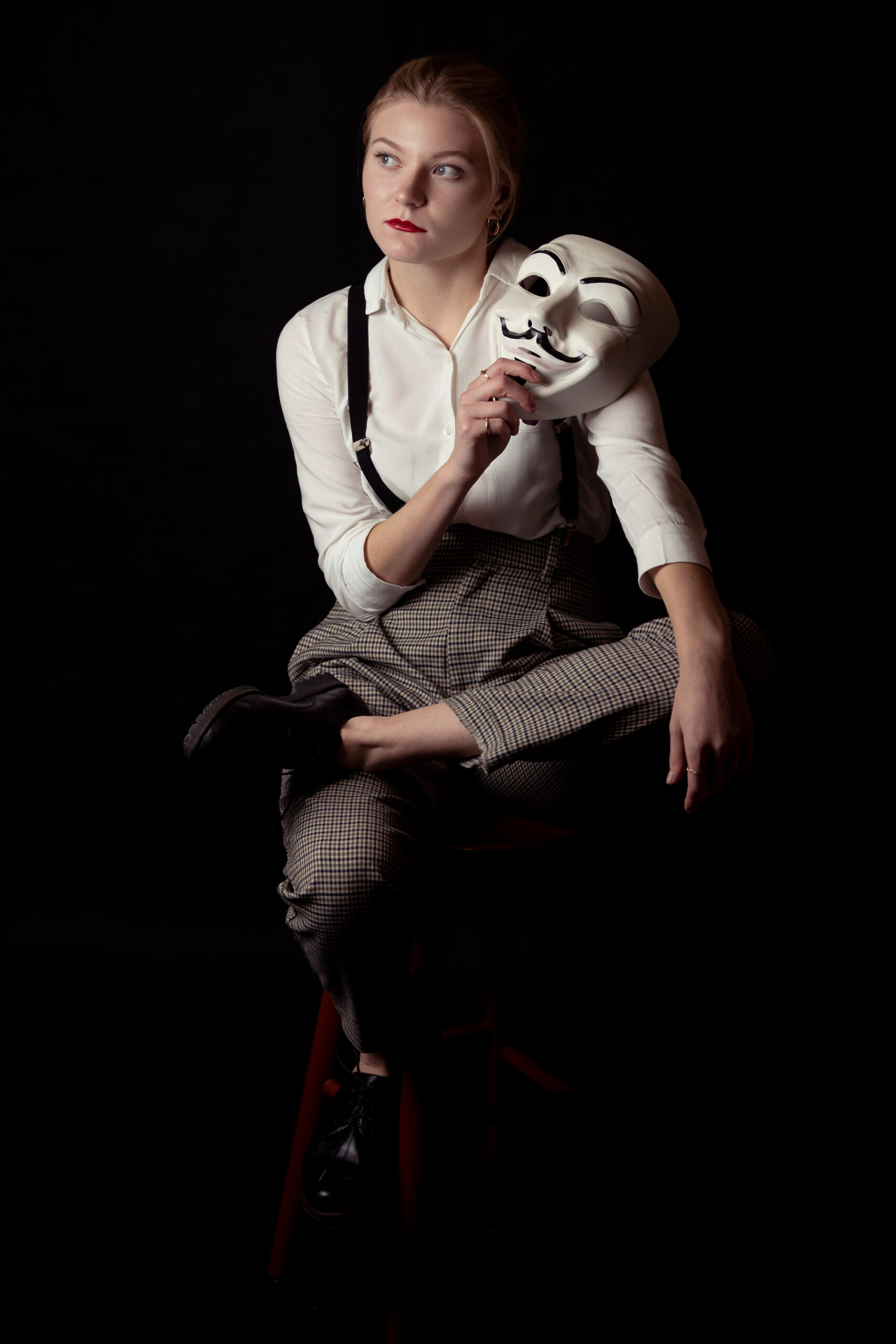 kobieta w białej koszuli, szelkach, spodniach w kratę, z czerwonymi ustami z maską i melonikiem w ręce
