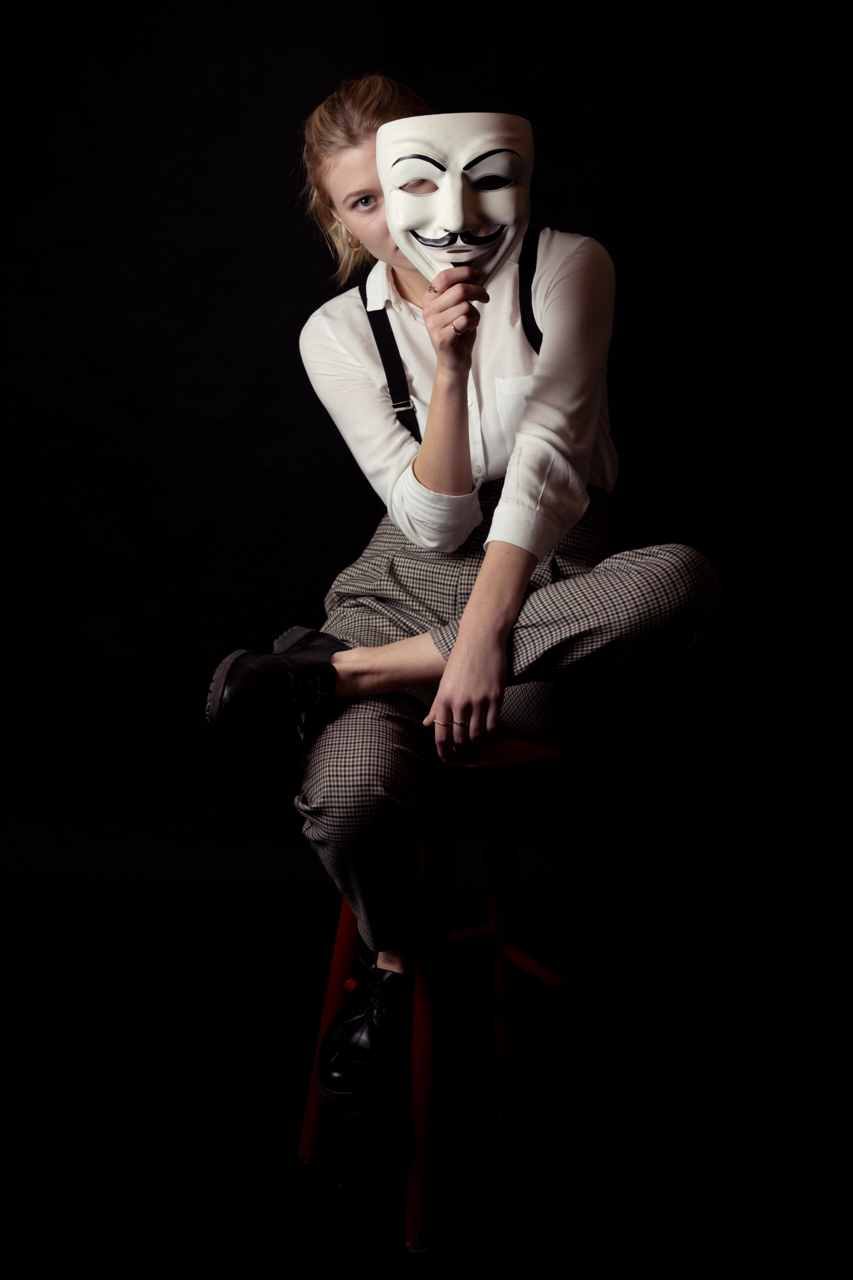 kobieta w białej koszuli, szelkach, spodniach w kratę, z czerwonymi ustami z maską i melonikiem w ręce